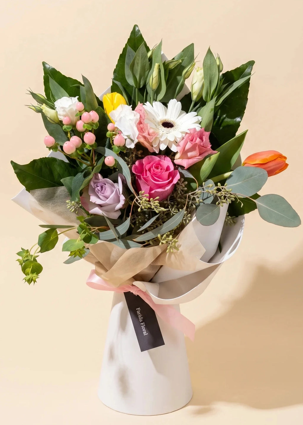 A beautifully arranged floral bouquet featuring pink roses, white daisies, a purple lavender, a yellow accent flower, and a bright orange tulip, wrapped in white and beige paper with a pink ribbon and a black tag, set against a solid beige background.