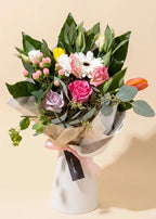 A beautifully arranged floral bouquet featuring pink roses, white daisies, a purple lavender, a yellow accent flower, and a bright orange tulip, wrapped in white and beige paper with a pink ribbon and a black tag, set against a solid beige background.